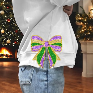 Op de afbeelding: Een wit sweatshirt met een grote paarse, groene en gouden glitter strik op de rug. De strik zit in het midden op de rug van het sweatshirt en is iets groter dan de hand van de drager.