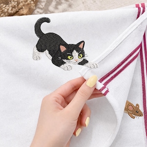 May include: White kitchen towel with an embroidered black and white cat stretching towards a brown mouse. The towel has red stripes along the edge.