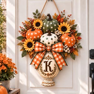 May include: A decorative door hanging featuring three pumpkins with white polka dots in green, white, and orange. The arrangement includes yellow sunflowers, orange berries, and autumn leaves, accented by a large orange and white gingham bow and a cream-colored urn base displaying the letter K.