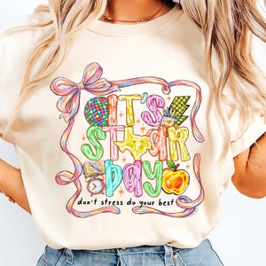 May include: Cream-colored t-shirt with a colorful graphic that reads "IT'S STAR DAY" with a disco ball, Texas shape, apple, and other school-themed elements. The text "don't stress do your best" is also included.