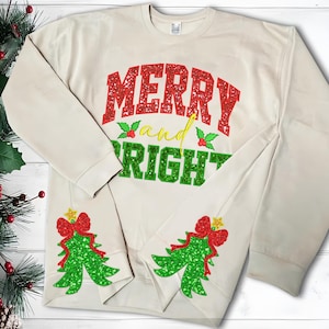 May include: Cream-colored sweatshirt with the words "MERRY and BRIGHT" in red and green glitter lettering. The sleeves feature green glitter Christmas trees with red bows and gold stars. A festive holiday design.