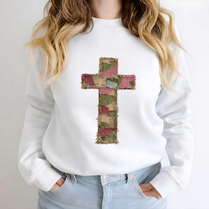 May include: White sweatshirt featuring a cross design. The cross is made of patchwork fabric in shades of green, pink, and tan. The sweatshirt has long sleeves and a crew neck. The overall style is casual and comfortable.