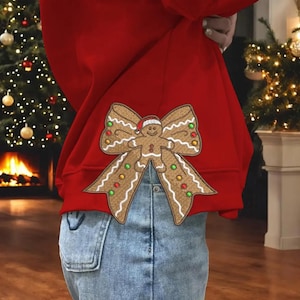 May include: A red sweatshirt with a gingerbread man bow design. The gingerbread man has a red hat and is surrounded by a bow with green and red accents. The sweatshirt is paired with blue jeans.