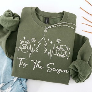 Op de afbeelding: Een olijfgroene sweatshirt met de tekst "Tis The Season" in witte letters. Het ontwerp bevat een rendier, kerstboom en sneeuwpop, verbonden door een hartslaglijn. Een zilveren ketting met een hanger ligt op de sweatshirt.