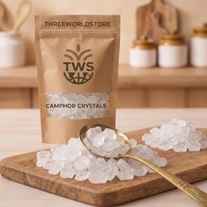 May include: A brown paper bag labeled "THREEWORLDSTORE" with a clear window displaying camphor crystals. A gold spoon and wooden cutting board hold more crystals. The bag reads "CAMPHOR CRYSTALS".