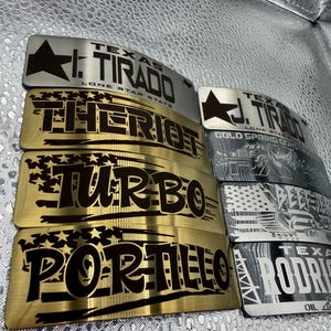 May include: A collection of rectangular metal nameplates in gold and silver finishes. Each plate features engraved black text including names like I. TIRADO, THERIOT, TURBO, PORTILLO, J. TIRADO, and RODRIGUEZ, alongside Texas-themed graphics and stars.