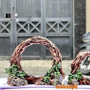 May include: Two decorative arches made of twisted brown vines with green plants growing from the base. The arches are arranged in a circle shape and are set on a white surface.