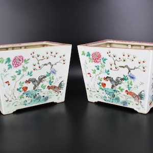 Puede incluir: Dos jardineras de cerámica blanca de forma rectangular y diseño ligeramente acampanado. Cada jardinera presenta un diseño pintado a mano de gallos, flores y ramas en tonos rojos, rosas, verdes y azules.