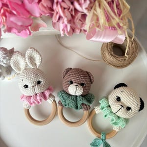 Pode incluir: Três chocalhos de bebê de crochê feitos à mão. Um coelhinho branco com babados rosa, um urso marrom com babados verdes e um panda com babados verdes. Cada animal é preso a um anel de madeira.
