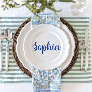 May include: A white ceramic plate with the name Sophia printed in blue script, centered on a white scalloped plate, a floral patterned napkin, and a round woven charger. Silverware includes two forks, two spoons, and a knife on a striped placemat.