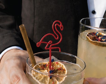 May include: A red flamingo-shaped cocktail stirrer made of wire, placed inside a glass filled with a light-colored beverage, ice, and a dehydrated citrus slice topped with small blue and red decorative sprinkles, alongside a brown straw.