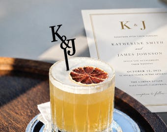 May include: A clear glass cocktail with a foamy top, garnished with a dried citrus slice and a black acrylic stirrer featuring the letters K&J. Beside it sits a wedding invitation for Katherine Smith and James Johnson.