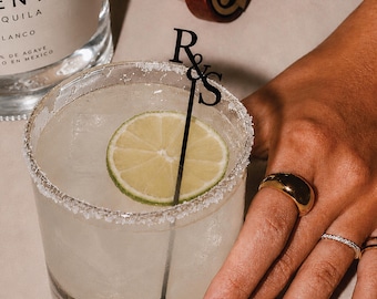 May include: A clear glass cocktail filled with a light-colored liquid, ice cubes, and a lime slice, featuring a black stirring stick with the letters R&S at the top. The rim of the glass is coated with salt, and a tequila bottle is visible nearby.