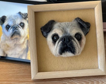 Puede incluir: Un retrato de un perro pug hecho con fieltro de aguja enmarcado. El pug tiene pelo negro alrededor de los ojos y la nariz, y el resto de su pelo es blanco y gris. El marco es de madera clara.