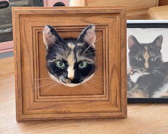Puede incluir: Un retrato de gato afieltrado tridimensional enmarcado. El gato tiene un patrón calicó con pelaje negro, canela y blanco y ojos verdes. El marco de madera es de color marrón cálido. Una fotografía del gato está al lado del retrato enmarcado.