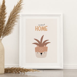 May include: A framed art print with a minimalist design. The artwork features a potted plant with a deer face, and the words "sweet HOME" in a playful font. The color palette includes beige, brown, and white tones. A vase with dried pampas grass is in the background.