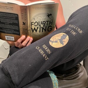 Könnte beinhalten: Eine Person liest ein Buch mit dem Titel "Fourth Wing" von Rebecca Yarros. Der Buchumschlag zeigt einen Drachen und die Worte "Fly or Die". Die Person trägt eine schwarze Jogginghose mit dem Aufdruck "Welcome to the Riders Quadrant" auf dem Bein.