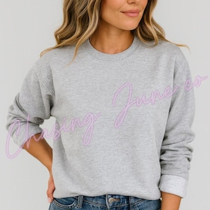 May include: A heather gray sweatshirt with the text "Chasing June co" in a light purple script. The sweatshirt has a crew neck and long sleeves with ribbed cuffs. The model is wearing blue jeans.