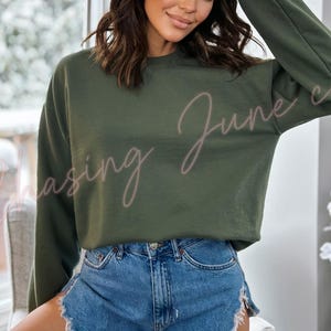 May include: An olive green sweatshirt with the text "Chasing June co" in a cursive font. The sweatshirt is paired with denim shorts. The image is taken indoors, with a window and floral arrangement in the background.