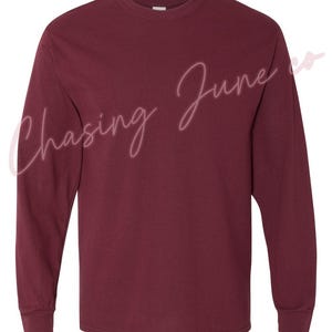 May include: A long-sleeved, crew neck t-shirt in a deep burgundy colour. The shirt has a classic fit with ribbed cuffs and a straight hem. The text "Chasing June co" is written in a cursive font across the front of the shirt.