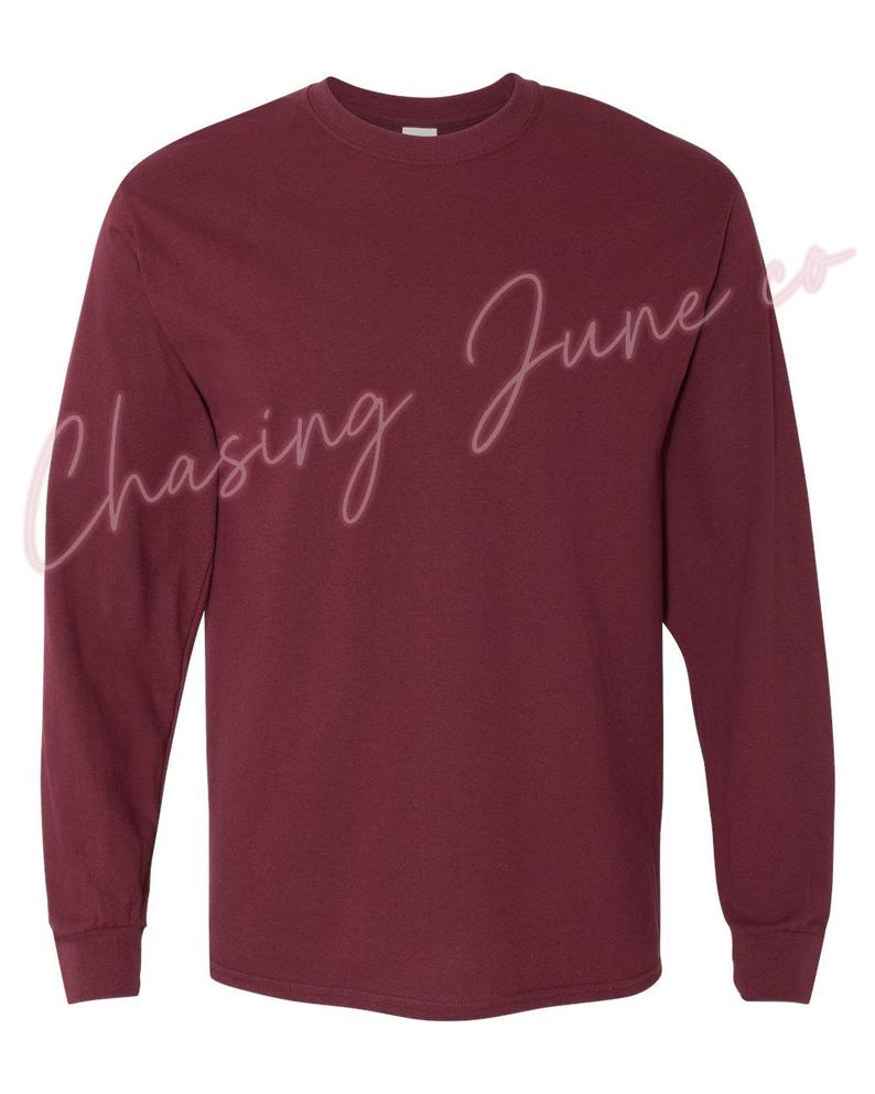 May include: A long-sleeved, crew neck t-shirt in a deep burgundy colour. The shirt has a classic fit with ribbed cuffs and a straight hem. The text "Chasing June co" is written in a cursive font across the front of the shirt.