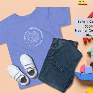 May include: A light blue t-shirt with a white graphic of a brain with the text "Not All Great Minds Think Alike" and "Embrace Neurodiversity". The shirt is folded and displayed with a pair of white sneakers and blue jeans.