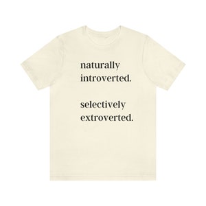 May include: Cream-colored t-shirt with the text "naturally introverted. selectively extroverted." in a simple black font. The shirt has short sleeves and a crew neck.