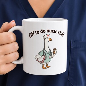 May include: A white ceramic mug featuring a cartoon illustration of a goose dressed as a nurse, wearing a cap with a red cross, a stethoscope, and holding a clipboard and a coffee cup. The text above the goose reads, Off to do nurse stuff.