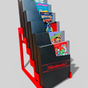 Pode incluir: Um suporte de exibição vermelho e preto para cartuchos de videogame com vários cartuchos de jogos Nintendo. O suporte tem um design em degraus e o logotipo da Nintendo é visível na frente do cartucho inferior.