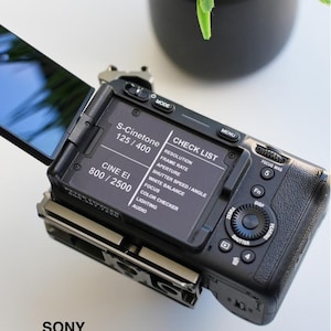 Puede incluir: Una cámara Sony FX30 negra con una pantalla abatible. La cámara tiene un menú de lista de verificación en la pantalla con opciones para la resolución, la velocidad de fotogramas, la apertura, la velocidad de obturación, el balance de blancos, el enfoque, el verificador de color, la iluminación y el audio. La cámara también tiene un dial para ajustar la configuración.