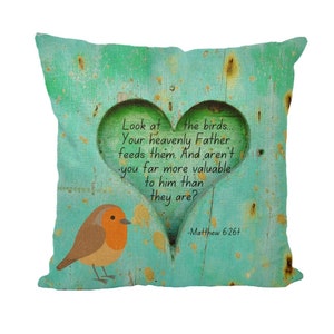 May include: A turquoise blue throw pillow with a distressed woodgrain pattern and a heart-shaped cutout. The cutout features a quote from Matthew 6:26+ about God's care for his children. A small robin bird is perched on the bottom left corner of the pillow.