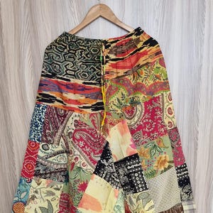 Peut inclure: Pantalon bouffant patchwork avec une variété de motifs de tissus colorés, dont des motifs cachemire, floraux et géométriques. Le pantalon est suspendu à un cintre en bois sur un fond en bois clair.