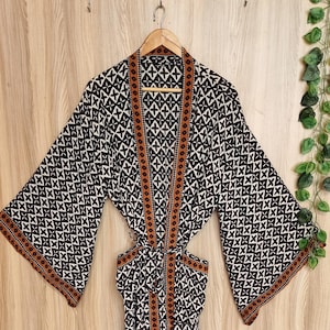 May include: A long-sleeved robe featuring a black and white geometric cross pattern throughout. The garment includes a brown border along the front opening and sleeve cuffs, with a matching tie belt at the waist to secure the front closure.
