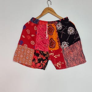 Puede incluir: Pantalones cortos de patchwork con una variedad de paneles de tela coloridos en tonos rojos, naranjas, negros y rosas. Los pantalones tienen una cintura elástica con un cordón blanco y muestran diversos patrones florales, geométricos y abstractos.