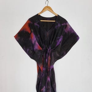 May include: A black kaftan dress featuring a v-neckline and a drawstring waist. The fabric displays a tie-dye pattern with blended streaks of deep purple and vibrant orange throughout the garment, hanging from a wooden hanger against a wall.