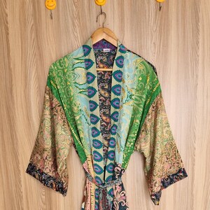 May include: A colorful, patterned kimono robe with a tie waist. The robe features a mix of green, blue, pink, and paisley patterns. It is hanging on a wooden hanger against a light wood background.