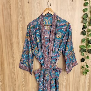 Puede incluir: Una bata con estampado de cachemira en color turquesa con cinturón a juego. La bata presenta un patrón detallado en tonos turquesa, morado y marrón. La bata cuelga sobre un fondo de madera, mostrando su diseño fluido y colores vibrantes.