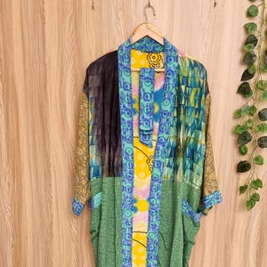 May include: A patchwork kimono robe in a variety of colours and patterns, including green, blue, yellow, and black. The robe is hanging on a wooden hanger against a wood-panelled wall.