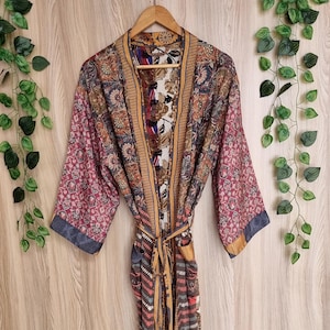 May include: A colorful, patterned kimono robe with a tie belt, hanging on a wooden hanger. The robe features a mix of floral and geometric designs in shades of pink, blue, brown, and gold. The sleeves have a darker trim.