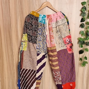 Può includere: Pantaloni patchwork multicolore con vari motivi, tra cui disegni floreali, a righe e paisley. I pantaloni sono appesi a una gruccia di legno su uno sfondo di legno chiaro. Hanno una vita con coulisse e caviglie arricciate.