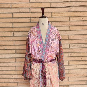 May include: A lightweight kimono-style robe featuring a vibrant mix of pink, peach, and deep red paisley patterns. The garment includes long sleeves, a V-neckline, and a matching fabric tie belt at the waist, displayed on a mannequin with the number 12.