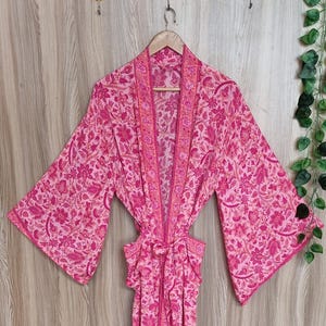 May include: A long-sleeved kimono robe featuring an all-over floral and paisley pattern in various shades of pink and magenta. The garment includes a matching fabric tie belt at the waist, wide sleeves, and a contrasting vertical border trim.