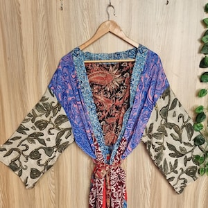 May include: A kimono-style wrap robe featuring a patchwork design with various patterns and colors including blue, red, black, and cream. The garment has wide sleeves, a tie-front closure, and a mix of floral and paisley prints throughout the fabric.