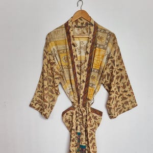 May include: A long-sleeved robe featuring a beige base with a repeating brown paisley pattern. The garment includes a contrasting dark trim along the front opening and a matching tie belt at the waist, with a vertical panel of purple and teal accents.