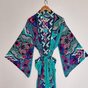 May include: A long-sleeved robe featuring a vibrant, multi-colored floral and geometric pattern in shades of teal, magenta, blue, and white. The garment includes a matching fabric tie belt at the waist and wide, flared sleeves, hanging on a hanger.
