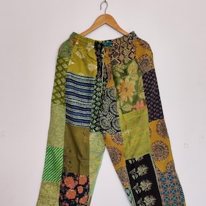 May include: Wide-leg trousers featuring a patchwork design with various fabric patterns in shades of olive green, mustard yellow, black, and blue. The trousers have an elastic waistband with a drawstring and are displayed on a wooden hanger.