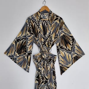 May include: A long-sleeved robe featuring a repeating art deco-inspired fan pattern in shades of black, gold, and slate blue. The garment has wide, flared sleeves, a wrap-front design with a matching tie belt, and a V-neckline, hanging on a wooden hanger.