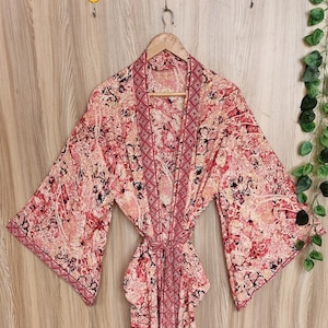 May include: A long-sleeved robe featuring a vibrant pink, red, and cream abstract floral pattern. The garment includes a matching fabric tie belt at the waist and a contrasting dark red geometric border along the front opening and sleeve cuffs.