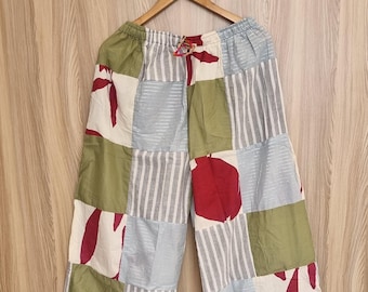 May include: Patchwork pants with a mix of colors including green, blue, white, and red. The pants are wide-legged and feature a variety of geometric patterns, including stripes and squares. They are hanging on a wooden hanger.