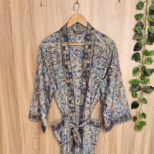 May include: A patterned robe with a paisley design in shades of blue, beige, and gold hangs on a wooden hanger. The robe has a tie belt and three-quarter sleeves. The background is a light wood panel.